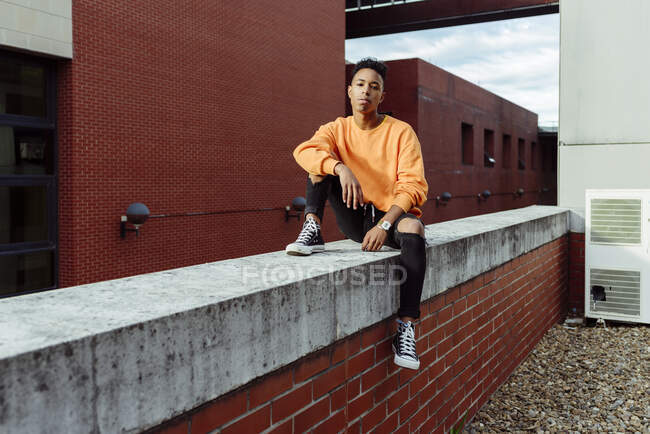 Joven adolescente étnico en ropa casual elegante sentado en la cerca del techo y mirando a la cámara - foto de stock