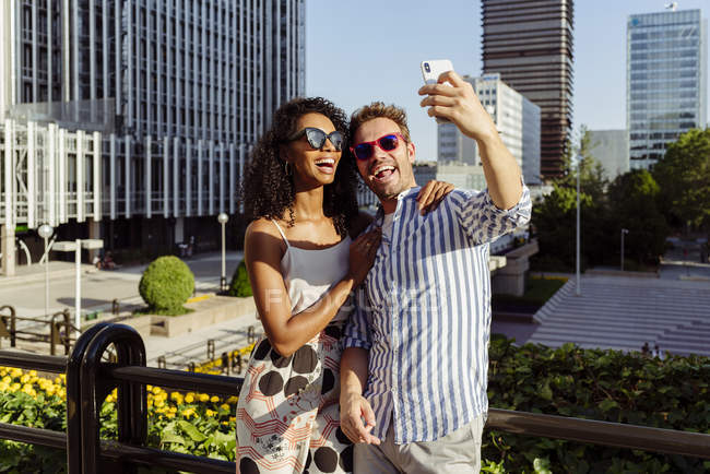 Couple multiracial joyeux posant pour selfie tout en se tenant sur le fond de la ville moderne — Photo de stock