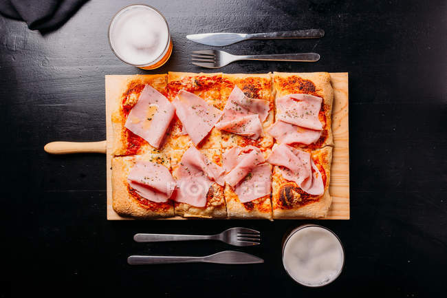 Pizza with sauce and slices of ham on wooden board on dark wooden table with drinks — Stock Photo