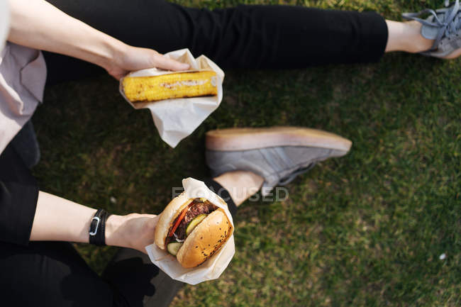 Woman sitting on grass in park holding takeaway burger and piece of cake — Stock Photo