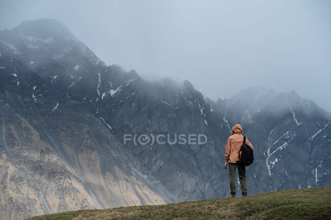 Vue arrière du mâle en vêtements chauds avec sac à dos randonnée dans les montagnes debout sur l'herbe regardant crête de montagne déchiquetée couverte de neige et des sommets cachés dans les nuages — Photo de stock
