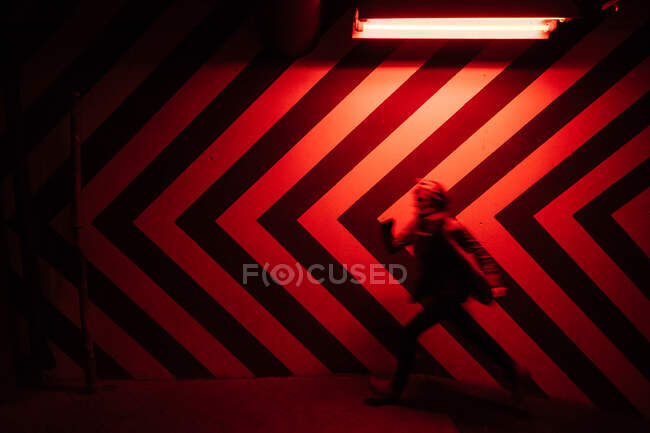 Vista lateral del movimiento figura borrosa del macho caminando por el túnel en dirección opuesta a las grandes flechas rojas y negras en la pared iluminadas por lámparas rojas - foto de stock