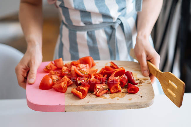 Primo piano delle mani femminili che tengono il tagliere con peperoni rossi tagliati a fette e pomodori in cucina — Foto stock