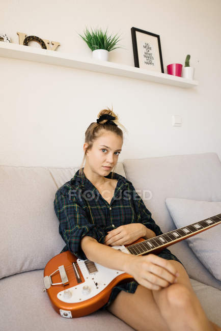 Retrato de mujer joven sosteniendo la guitarra eléctrica mientras está sentado en un cómodo sofá en la sala de estar - foto de stock