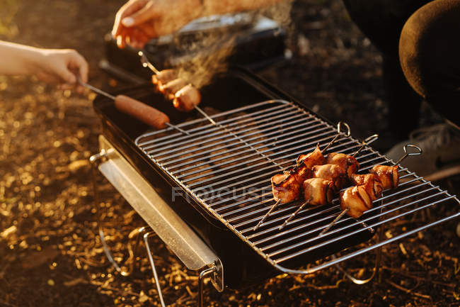 Mãos humanas preparando bacon e salsichas em espetos grelhando em carvão em chamas em grelha portátil ao ar livre — Fotografia de Stock