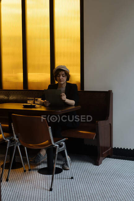 Junge hübsche Frau mit Mütze sitzt allein am Tisch in einem modernen Restaurant und liest sich durch die Speisekarte — Stockfoto