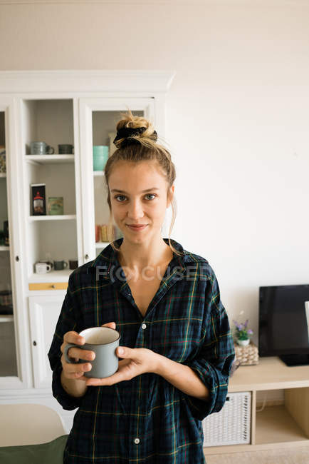 Retrato de jovem mulher em xadrez camisa de pé com caneca de café em casa — Fotografia de Stock