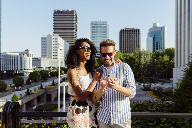 Смеющаяся многорасовая пара с помощью смартфона, стоя на фоне современного города — стоковое фото