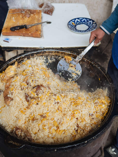 Tiro de colheita de cima do homem que prepara o prato nacional do Uzbequistão pilaf no enorme caldeirão metálico — Fotografia de Stock