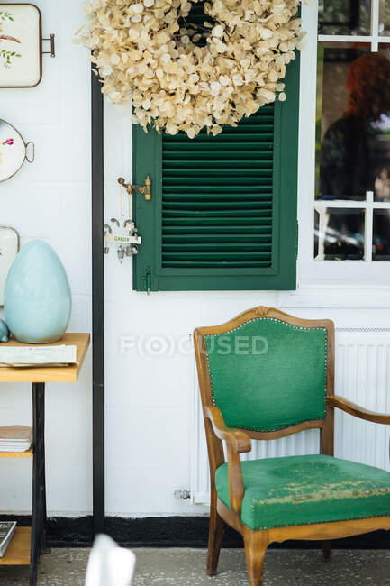 Elegante porche rural con persianas verdes con letrero de jardín creativo y mesa de madera con silla de tapicería antigua y corona de pétalos de planta seca colgando por encima - foto de stock