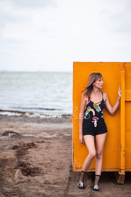 Slender woman in shorts and top leaning on orange metal wall while standing on sandy beach at seaside — Stock Photo