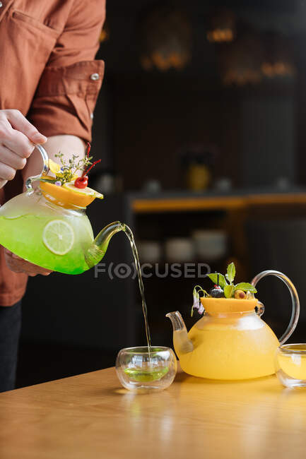Crop man serving creative cocktail from glass pot with fruit on top into small glass cup on table — Stock Photo