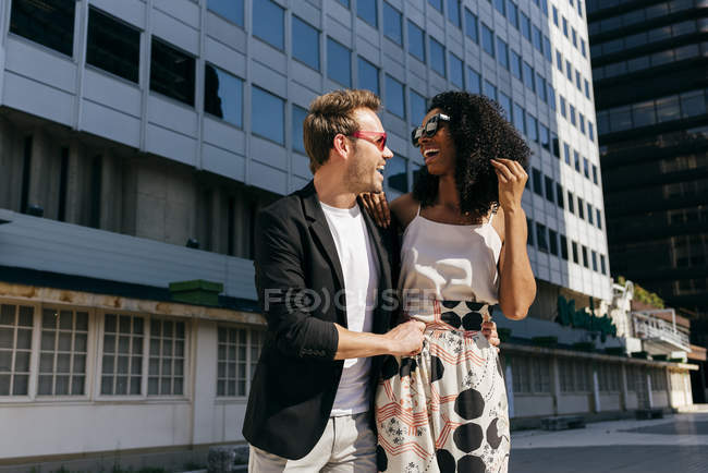 Riéndose pareja multirracial en gafas de sol caminando juntos en la calle de la ciudad - foto de stock