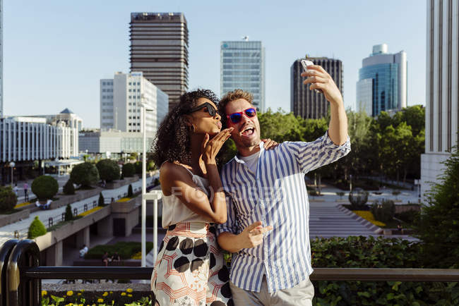 Cheerful multiracial couple posing for selfie while standing on background  of modern city — making faces, daytime - Stock Photo | #214402192