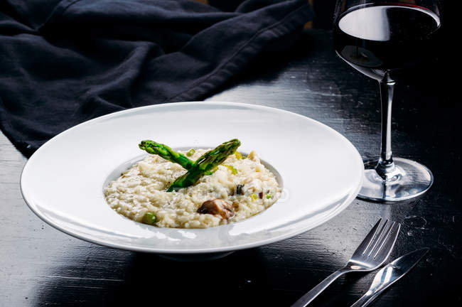 Вегетарианское ризотто и овощи на белой фарфоровой тарелке на темном столе с вилкой, ножом и бокалом вина — стоковое фото