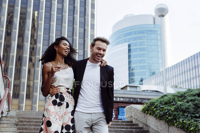 Riendo elegante pareja multirracial caminando escaleras abajo juntos en la calle de la ciudad - foto de stock