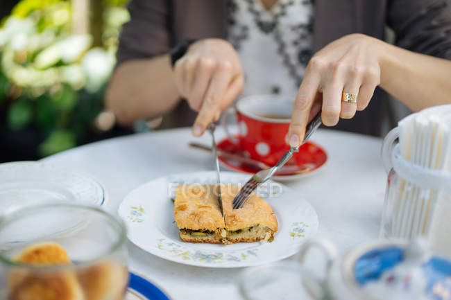 Agujas femeninas cortando trozo de pasta en plato con cuchillo y tenedor en mesa de jardín - foto de stock