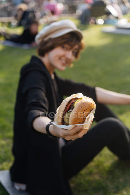 Sorrindo mulher mostrando hambúrguer enquanto sentado na grama no parque — Fotografia de Stock