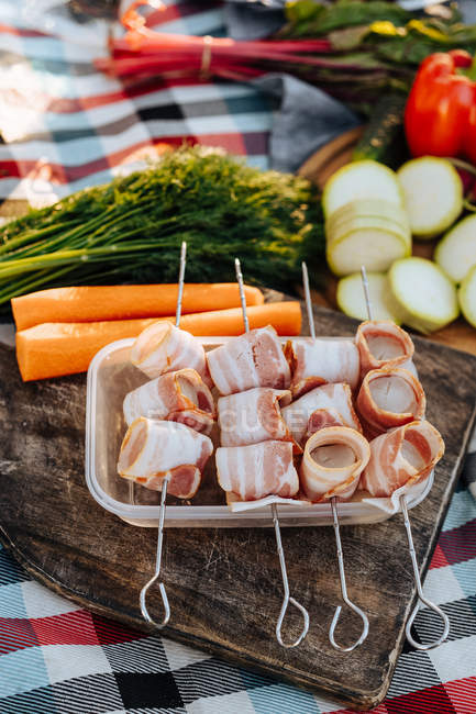 Close-up of metal skewers with strips of bacon on dish on wooden board — Stock Photo