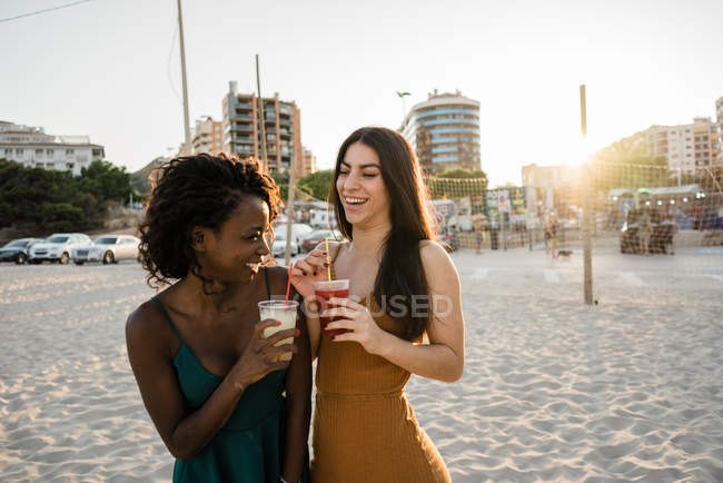Young women chatting and laughing with drinks on sandy town beach — Stock Photo