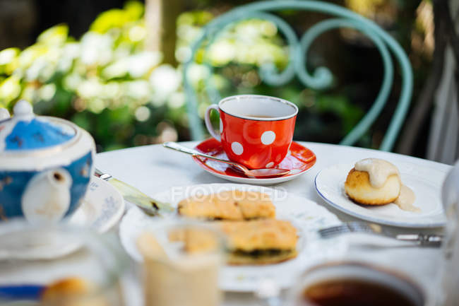 Red ceramic polka-dotted mug on saucer and pieces of pasty on plate on garden table — Stock Photo