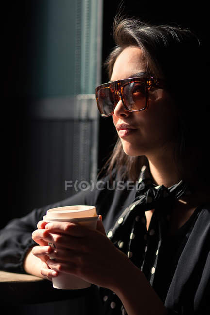 Mujer elegante en gafas de sol y pañuelo sosteniendo taza de papel de café y apoyándose en la mesa - foto de stock