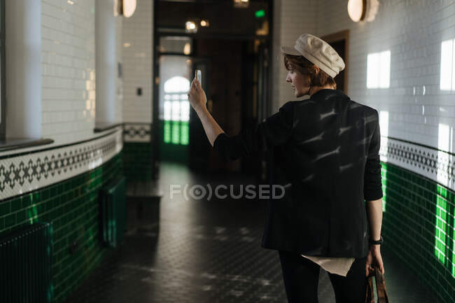 Vista trasera de la mujer joven de moda usando el teléfono y tomando selfie en el pasillo moderno de azulejos con lámparas - foto de stock