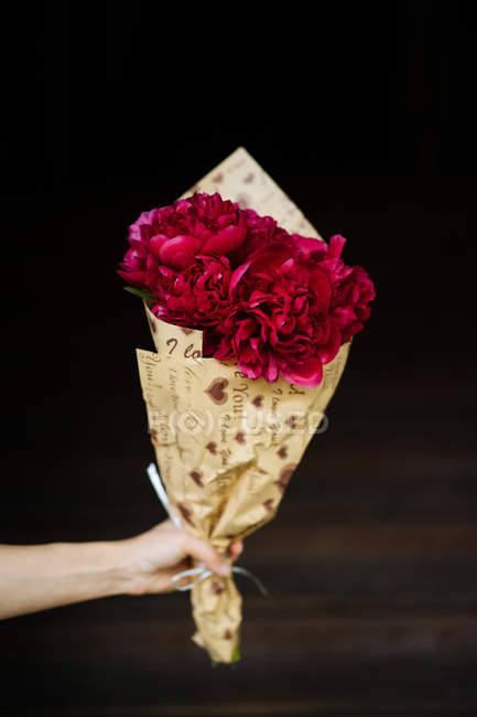 Mano umana che tiene bouquet di peonie rosa in carta da imballaggio su sfondo scuro — Foto stock