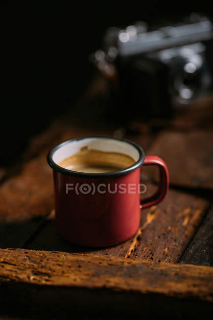Enamel cup of coffee on rustic wooden surface with retro camera on background — Stock Photo