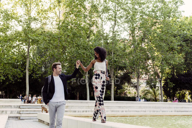 Очаровательная черная женщина улыбается и ходит по границе фонтана с парнем в парке — стоковое фото