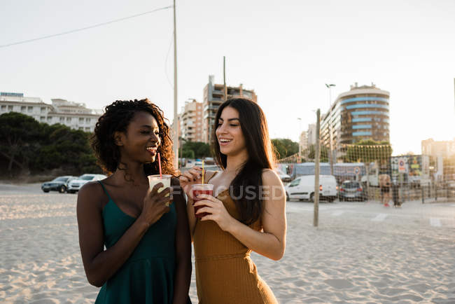 Young women chatting and laughing with drinks on sandy town beach — Stock Photo