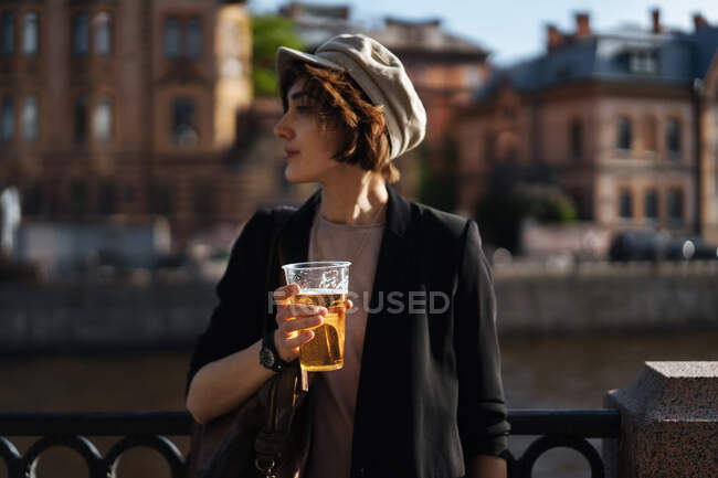 Side view of stylish girl in cap holding plastic cup with beer and standing on city embankment looking away in sunlight — Stock Photo