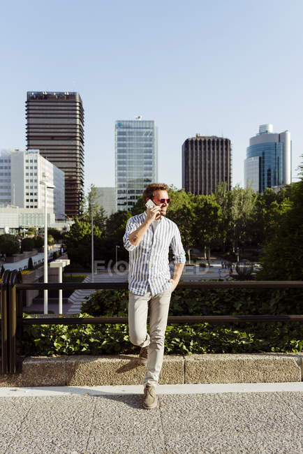 Элегантный мужчина, опирающийся на забор и разговаривающий на смартфоне в современном городе — стоковое фото