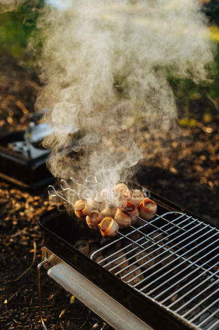 Primo piano della griglia portatile con carbone ardente e spiedini con strisce di pancetta grigliate — Foto stock