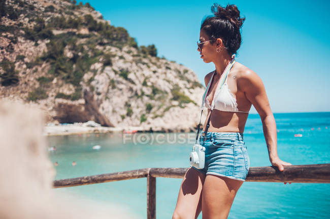 Niña en ropa de verano apoyada en pasamanos de madera en la playa - foto de stock
