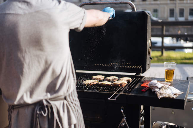 Man seasoning burger patties with cheese while grilling on griddle outdoors on street in sunlight — Stock Photo