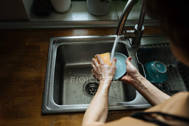 Crop back view of woman hands?cleaning blue saucer with dish-washing sponge in sink on wooden?counter with sill with pots nearby — Foto stock
