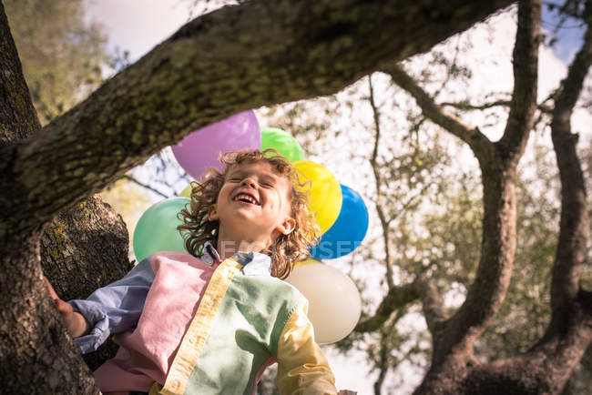 Low angle view of boy with eyes closed sitting on tree with balloons — Stock Photo