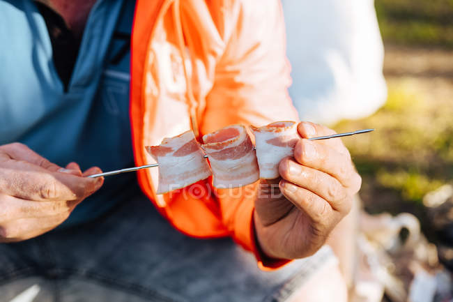 Human hands arranging folded bacon strips on metal skewer for barbecue meal — Stock Photo