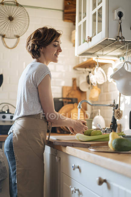 Женщина моет фрукты и овощи в раковине под потоком пресной воды на кухне — стоковое фото