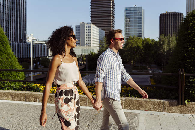 Sorrindo elegante casal multirracial andando de mãos dadas na cidade moderna — Fotografia de Stock