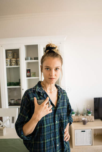 Retrato de mujer joven en camisa a cuadros de pie con taza de café en casa - foto de stock