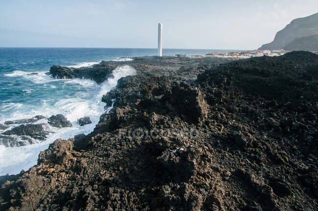 Picturesque view of gray rocky cliffs with tower of lighthouse on background on shoreline of ocean with strong waves — Stock Photo