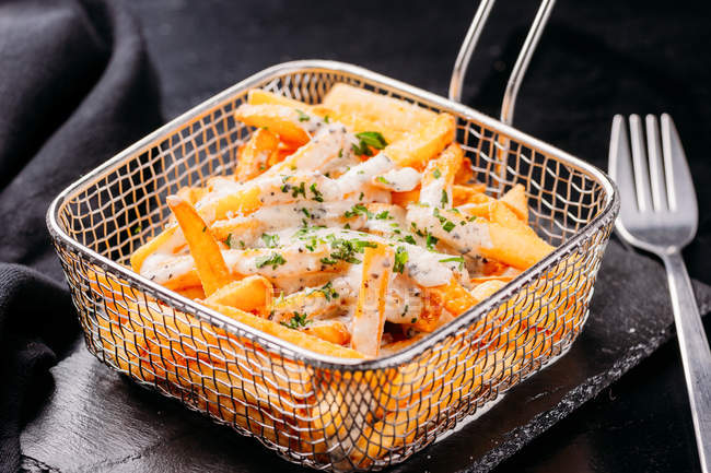 Fresh delicious French fries sprinkled with chopped potherbs with sauce in wire fryer basket on slate — Stock Photo