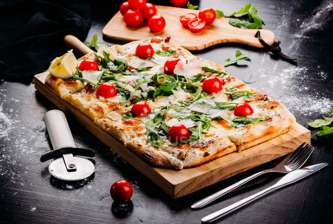 Pizza coupée rectangulaire aux tomates cerises, potherbs et fromage sur plateau en bois sur table noire — Photo de stock