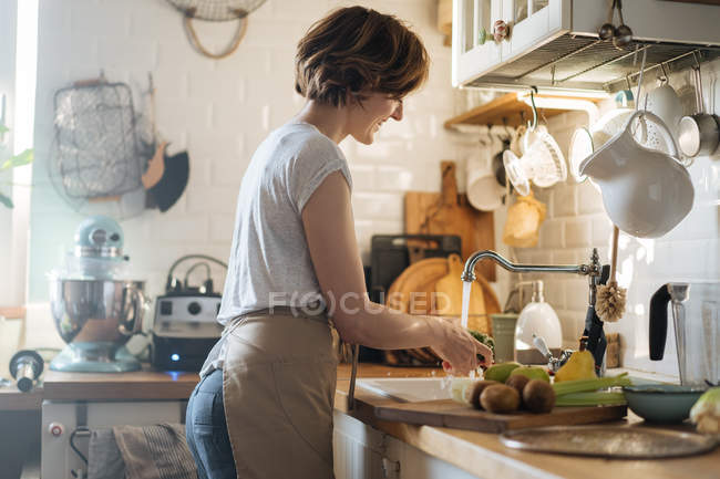 Mujer lavando frutas y verduras en fregadero bajo la corriente de agua dulce en la cocina - foto de stock