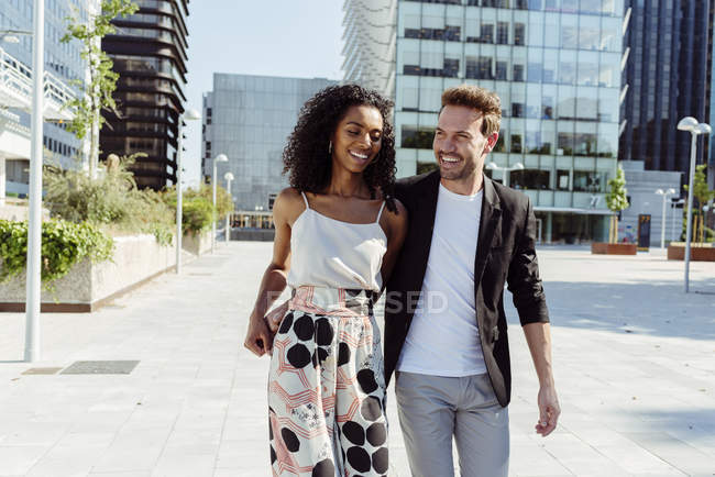 Lächelndes multiethnisches Paar hält Händchen, während es an einem sonnigen Tag gemeinsam auf der Stadtstraße spaziert — Stockfoto