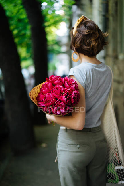 Rückansicht einer Frau in lässiger Sommerkleidung mit Vintage-Frisur und Accessoires, die auf dem Bürgersteig steht, mit leuchtend rosa auffälligen Blumen, die in Papier gehüllt sind, mit Bäumen auf verschwommenem Hintergrund — Stockfoto