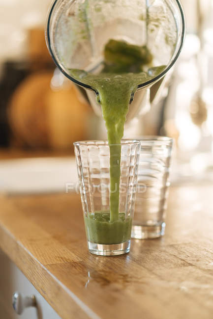 Versando sano frullato verde dalla tazza del frullatore in vetro sul bancone in legno in cucina — Foto stock