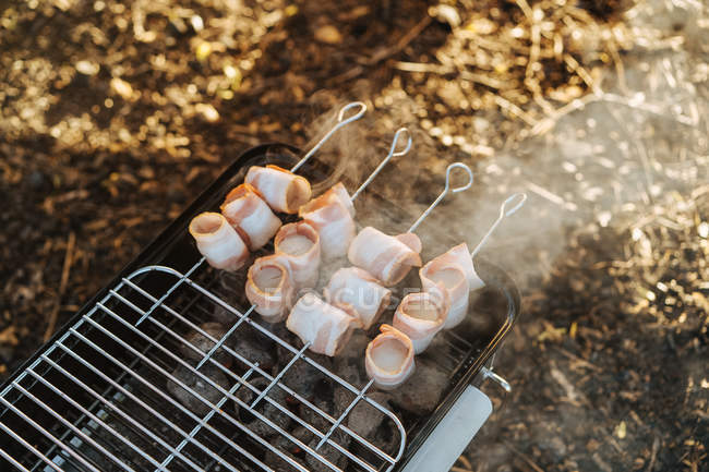 Close-up of portable griddle with burning charcoal and skewers with bacon strips grilling — Stock Photo